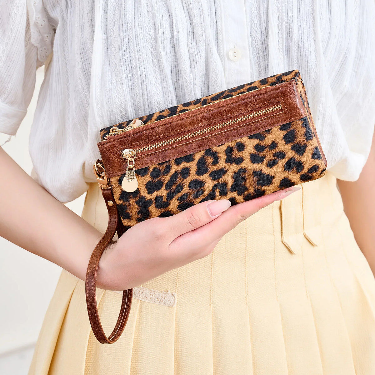 A hand holds a brown leopard print clutch wallet with a gold wristlet strap against a light yellow pleated skirt.