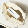 A beige canvas shoulder tote bag with light beige straps and a white rectangular item inside.