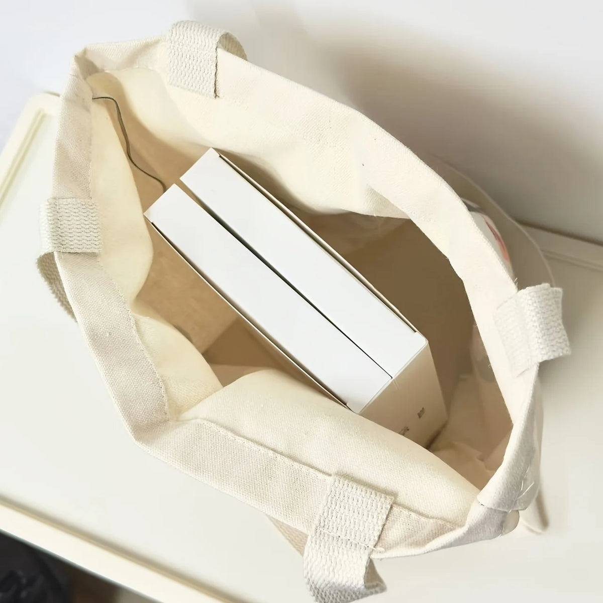 A beige canvas shoulder tote bag with light beige straps and a white rectangular item inside.