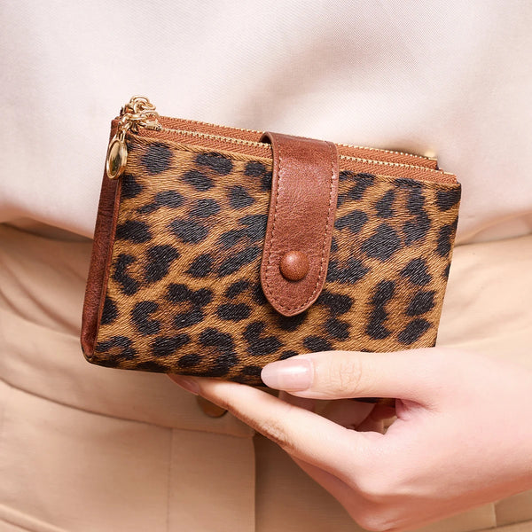 A brown leopard print women's wallet with a leather flap and gold-toned zipper is held in a hand.