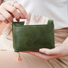 A hand holds a dark olive green minimalist foldable wallet with a zipper and a banknote.