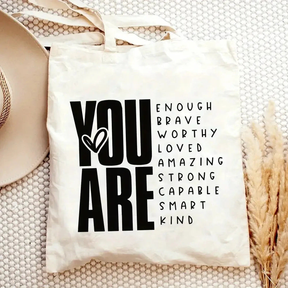 A white canvas tote bag with black text that reads YOU ARE ENOUGH BRAVE WORTHY LOVED AMAZING STRONG CAPABLE SMART KIND