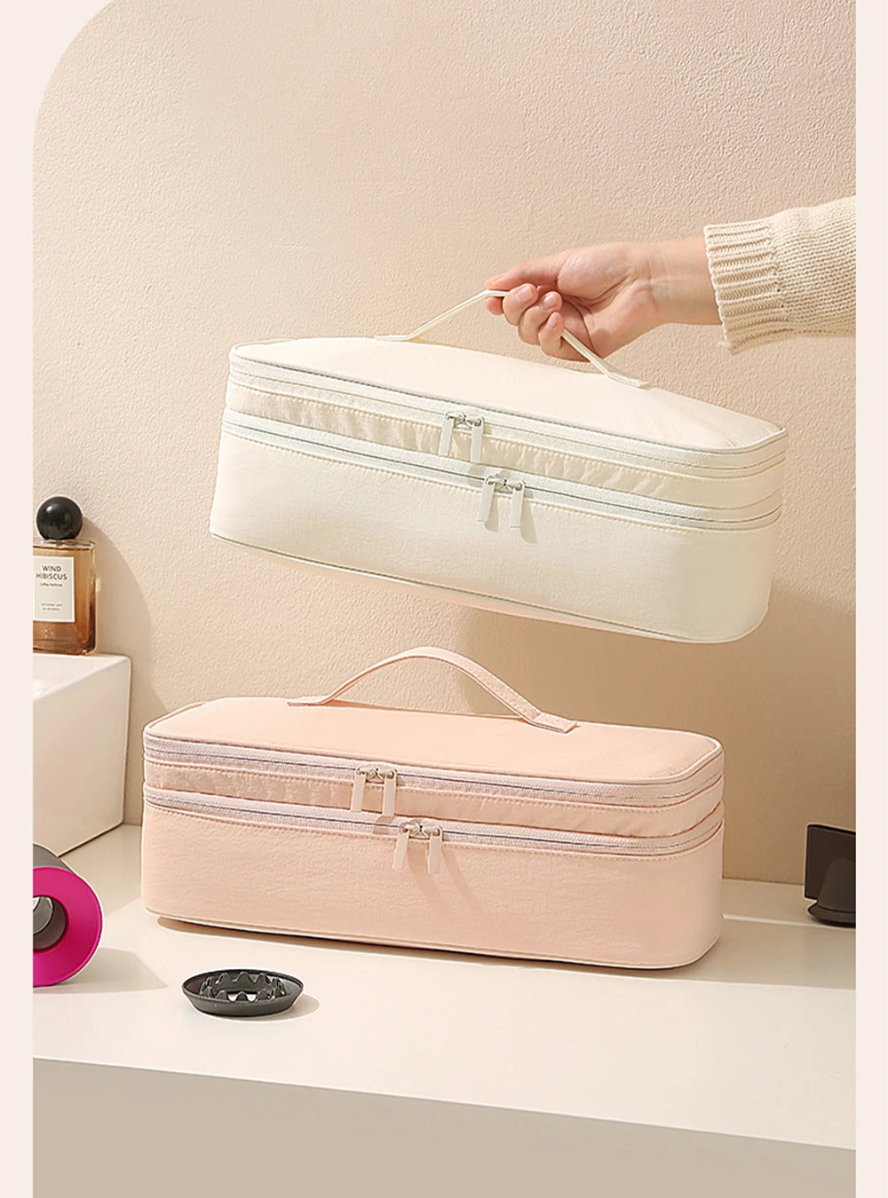 A hand is lifting a cream-colored storage bag for a hair dryer, with a pink version resting on a white surface.