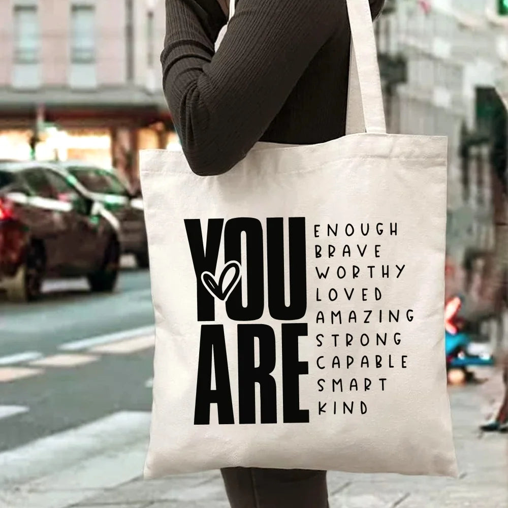 A white canvas tote bag with black text that reads YOU ARE ENOUGH BRAVE WORTHY LOVED AMAZING STRONG CAPABLE SMART KIND
