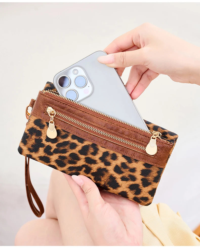 A hand holds a brown leopard print clutch wallet with a zipper and a wristlet strap, displaying a smartphone.
