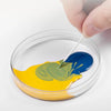 Une main applique une goutte de peinture bleue sur une surface jaune dans un récipient transparent.