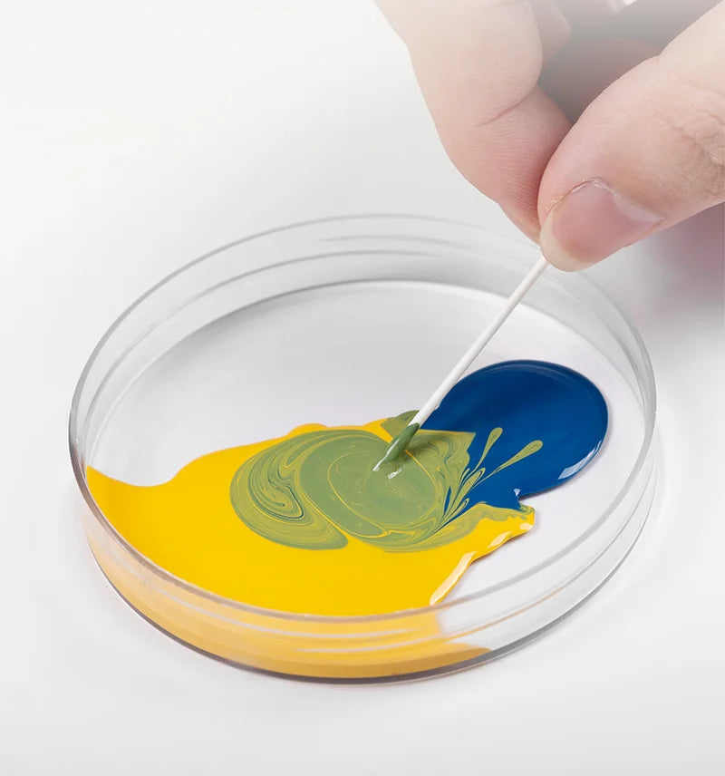 Une main applique une goutte de peinture bleue sur une surface jaune dans un récipient transparent.