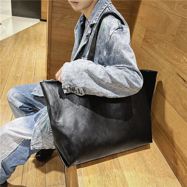 A person wearing a denim jacket is sitting on the floor next to an oversized black PU leather tote bag.