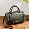 Dunkelolivgrbare Leder-Schultertasche mit Reißverschluss und rosaförmigem Anhänger auf Holzoberfläche.