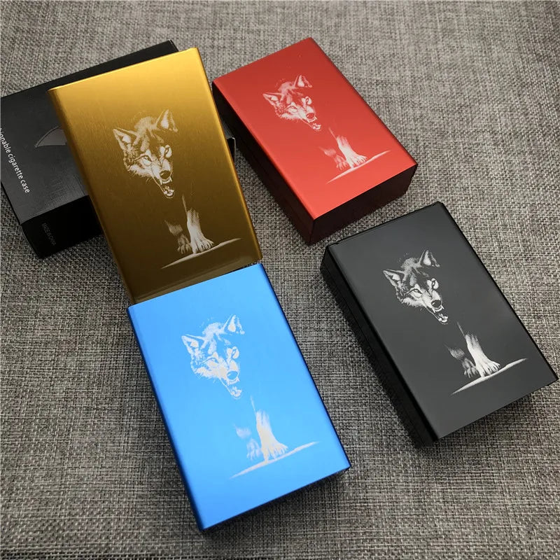 Three custom engraved cigarette cases in gold, red, and blue are displayed on a gray fabric surface.