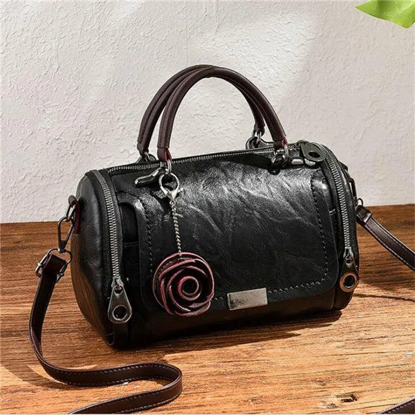 Schwarze Lederhandtasche mit braunem Riemen und pinkem Blumenanhänger auf Holzoberfläche.