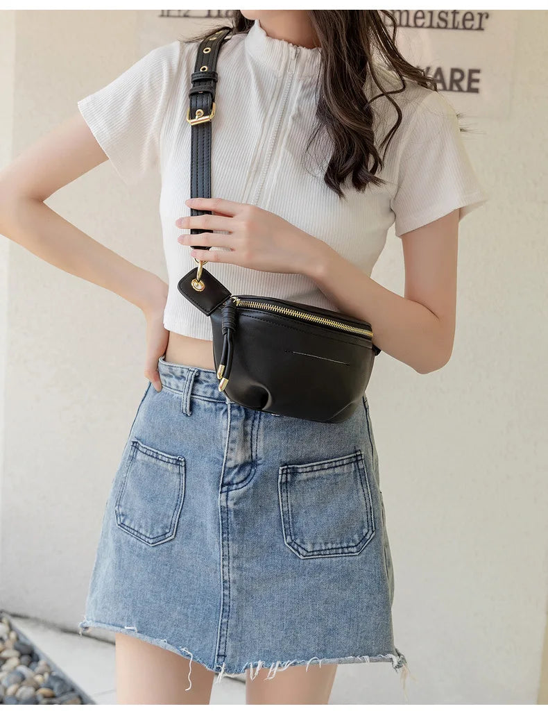 A woman models a black PU leather crossbody sling bag with a gold chain strap and a white top and denim skirt.