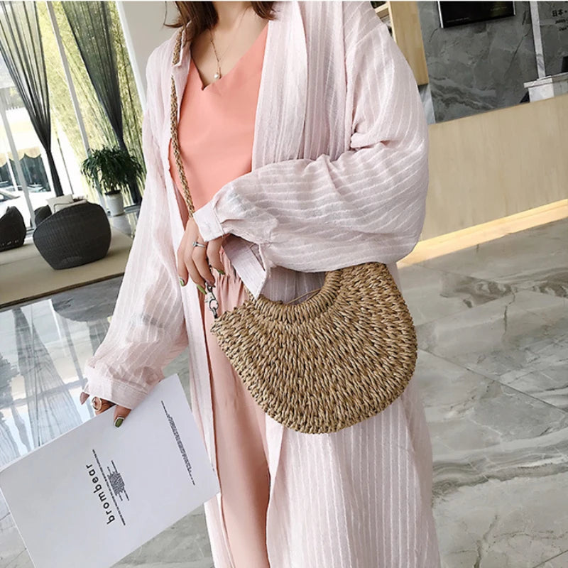 A woman holds a handwoven straw moon bag with a top handle in a light pink cardigan.
