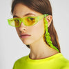 A close-up of a model wearing bright lime green acrylic sunglasses with a chunky yellow chain strap.