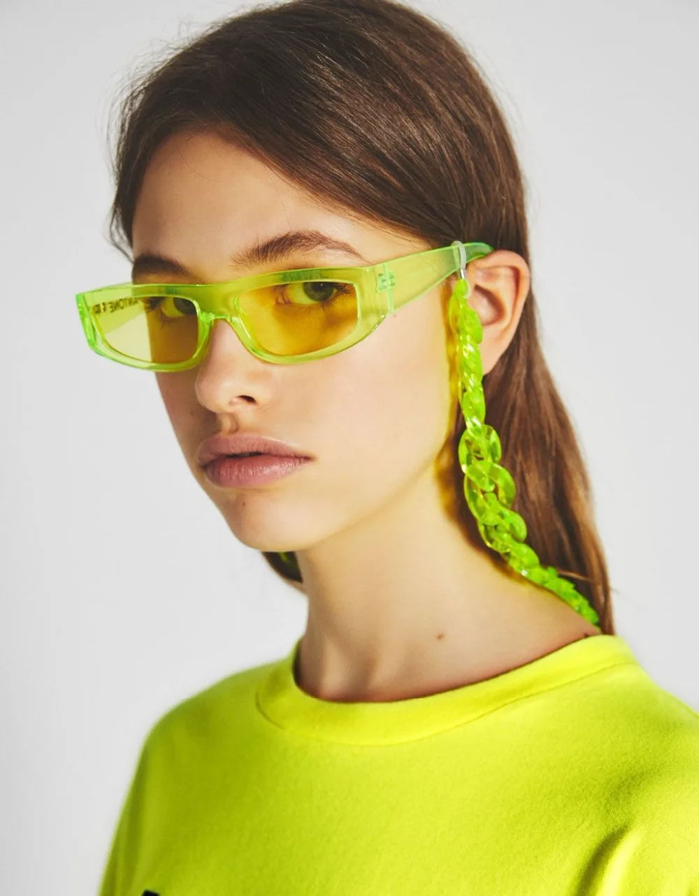 A close-up of a model wearing bright lime green acrylic sunglasses with a chunky yellow chain strap.