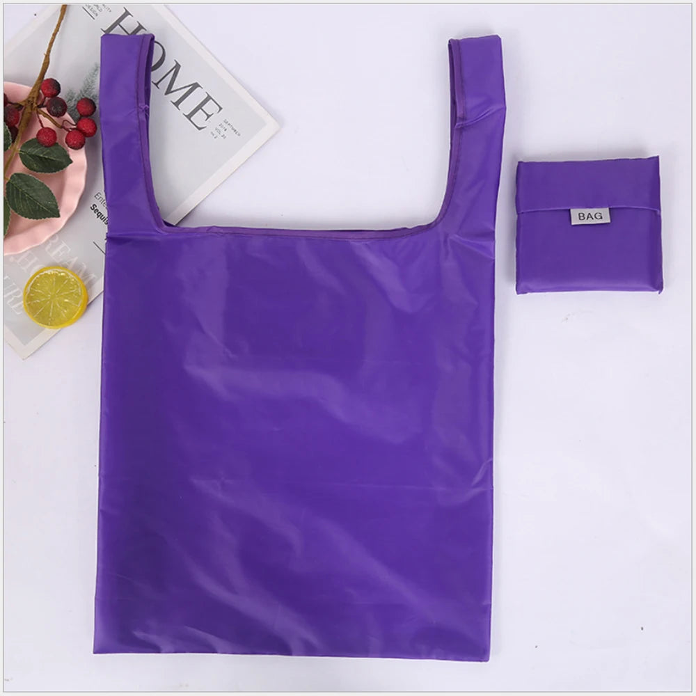 A purple, glossy, foldable shopping bag is displayed next to a small purple bag tag and some fruit.