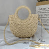 A light beige handwoven straw moon bag with a top handle and long strap is displayed on a white surface.