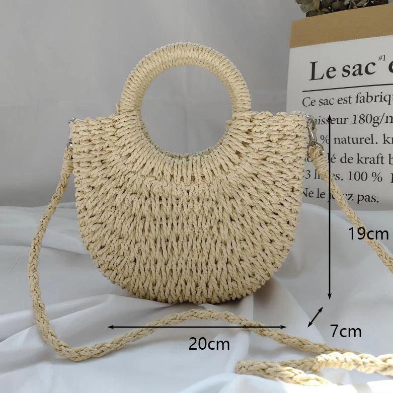 A light beige handwoven straw moon bag with a top handle and long strap is displayed on a white surface.