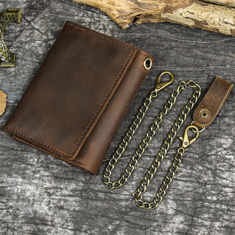 A brown genuine leather trifold wallet with a detachable gold-toned chain lying on a gray textured surface.