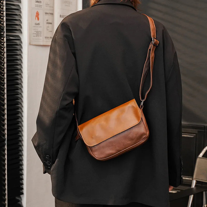 A person wearing a black blazer carries a brown leather crossbody bag on their shoulder.