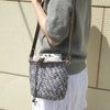 A hand wearing a grey sweater holds a woven gray bag with a leather crossbody strap.