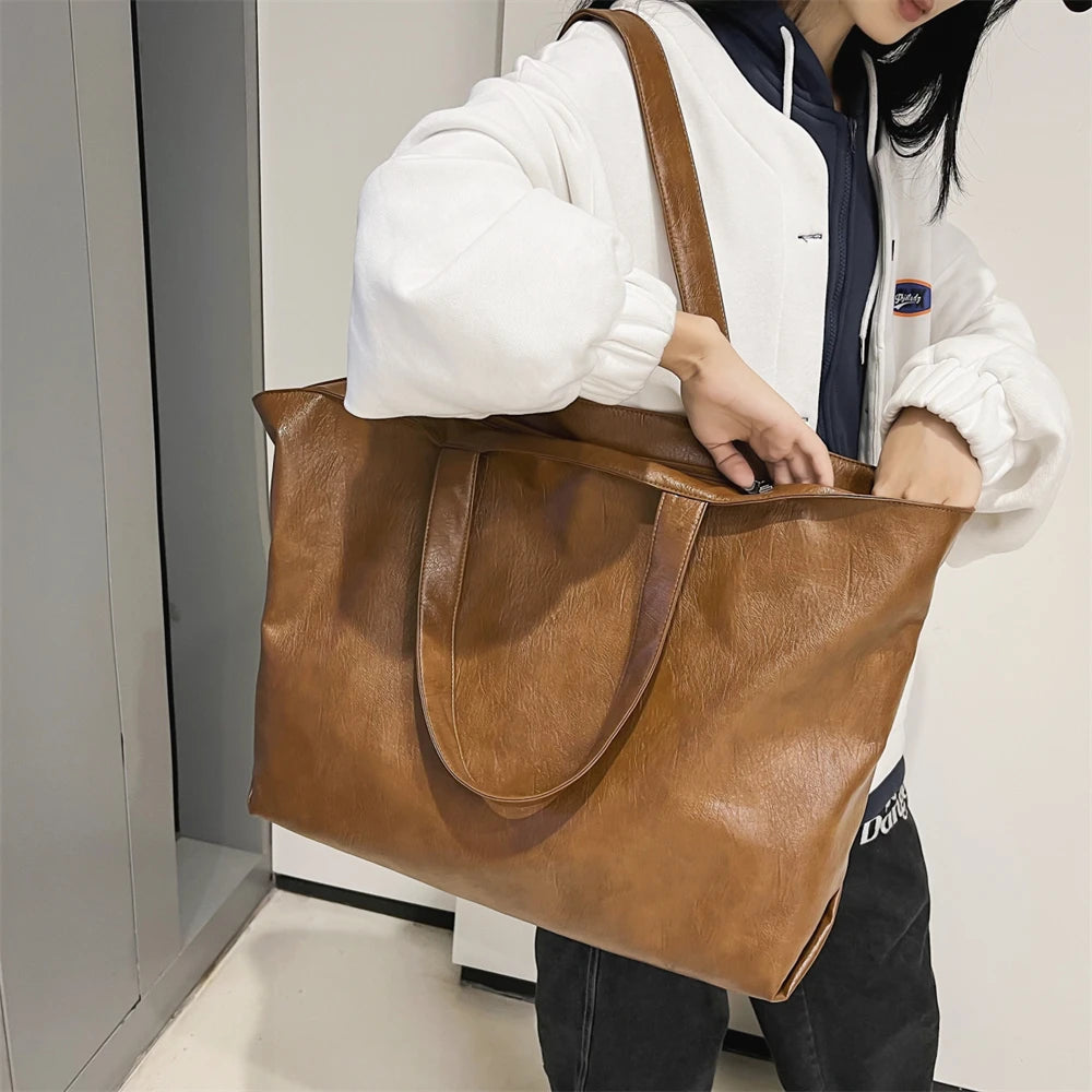 A person is holding a large brown leather tote bag with a zipper closure and a white hoodie.