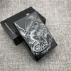 A black cigarette case with a custom engraved design of a dragon on the lid, resting on a gray fabric surface.