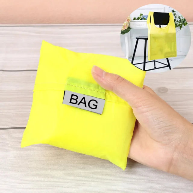 Une main tient un sac de shopping jaune vif et pliable écologique avec une étiquette noire sur une surface en bois clair.