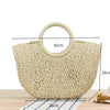 A beige handwoven straw moon bag with a top handle resting on a wooden surface.