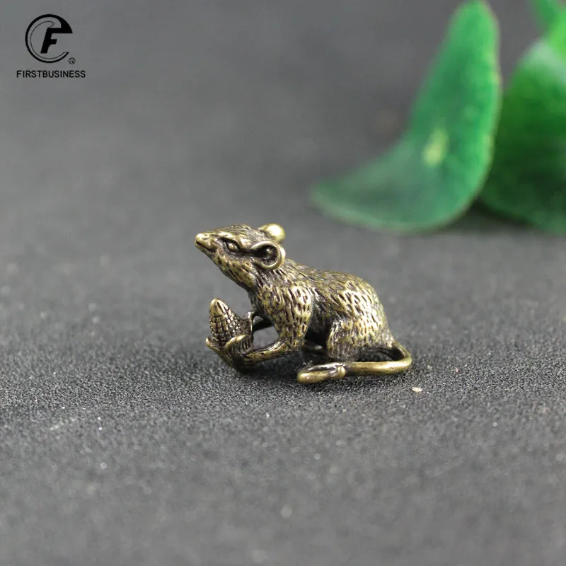 A vintage brass figurine of a rat is placed on a dark surface next to green leaves.