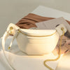 A light beige PU leather crossbody sling bag with gold hardware resting on a white surface next to a brown fabric.