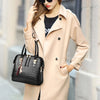 A woman wearing a beige trench coat holds a black crocodile-embossed leather tassel shoulder bag.