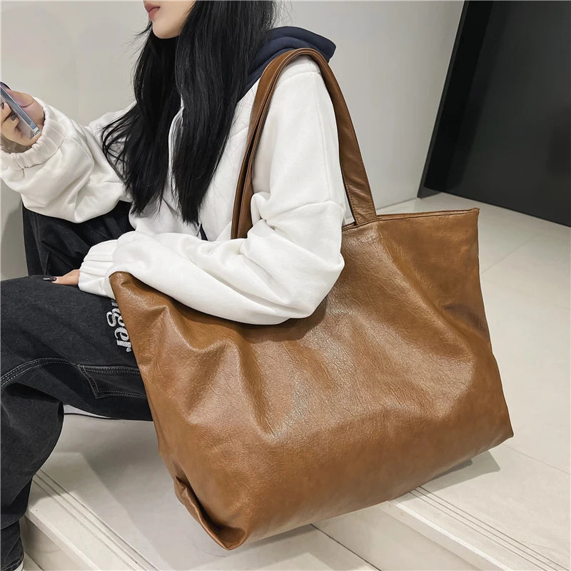 A woman holds an oversized brown PU leather tote bag with a zipper closure.