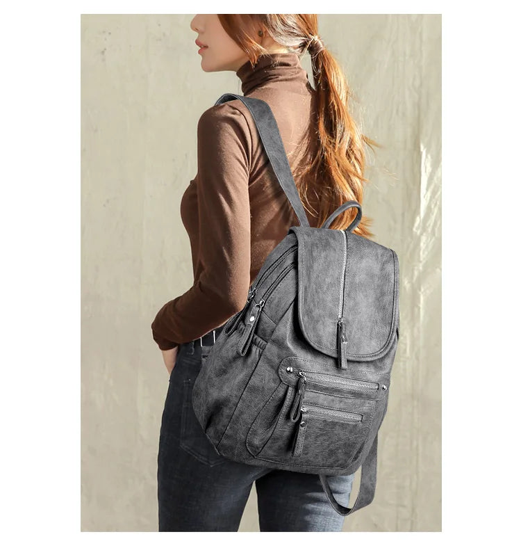 A woman wearing a brown turtleneck and dark jeans is shown carrying a gray vintage soft leather backpack.