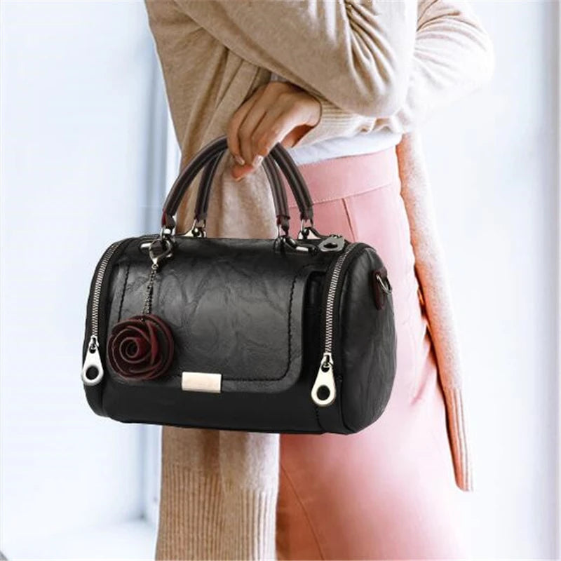 Eine Frau hält eine schwarze Leder-Blumen-Anhänger-Schultertasche vor einem hellen Hintergrund.