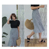 A woman models a handwoven straw moon bag with a top handle while wearing a black top and plaid skirt.