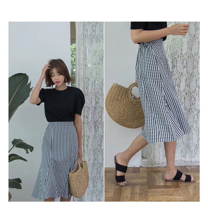 A woman models a handwoven straw moon bag with a top handle while wearing a black top and plaid skirt.