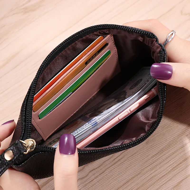 Una mano abre una cartera con cremallera de cuero sintético marrón oscuro, revelando varias tarjetas y un smartphone dentro.