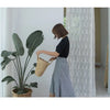 A woman in a black top and plaid skirt is holding a handwoven straw moon bag with a top handle.