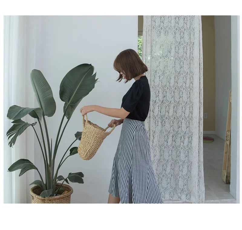 A woman in a black top and plaid skirt is holding a handwoven straw moon bag with a top handle.