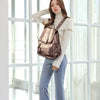 A woman wearing a beige turtleneck sweater and blue pants stands in front of a window holding a brown leather backpack.
