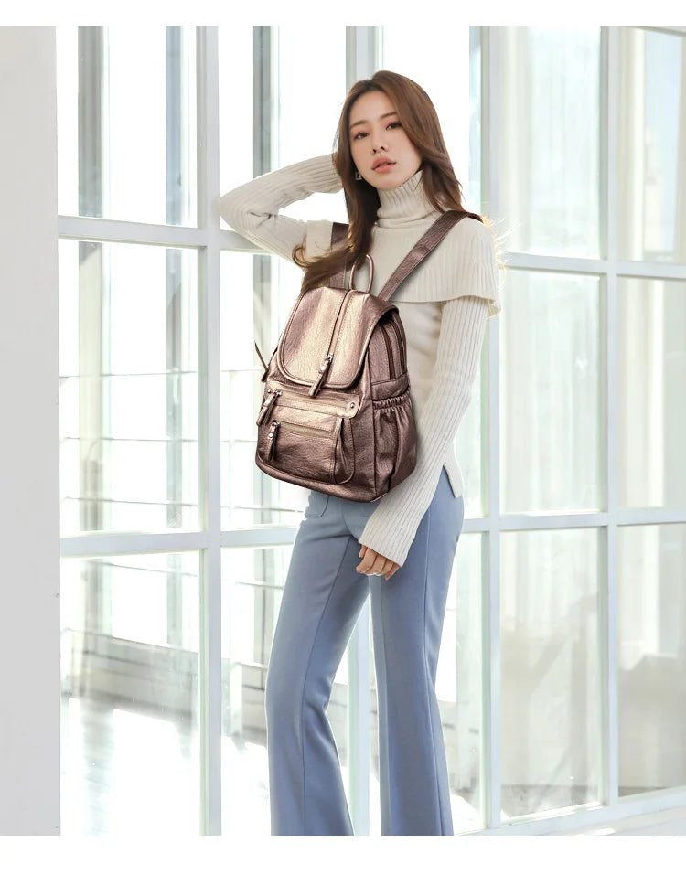 A woman wearing a beige turtleneck sweater and blue pants stands in front of a window holding a brown leather backpack.