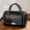 Schwarze Leder-Schultertasche mit braunem Henkel und rosem Blumenanhänger auf Holzoberfläche.