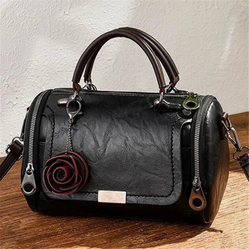 Schwarze Leder-Schultertasche mit braunem Henkel und rosem Blumenanhänger auf Holzoberfläche.