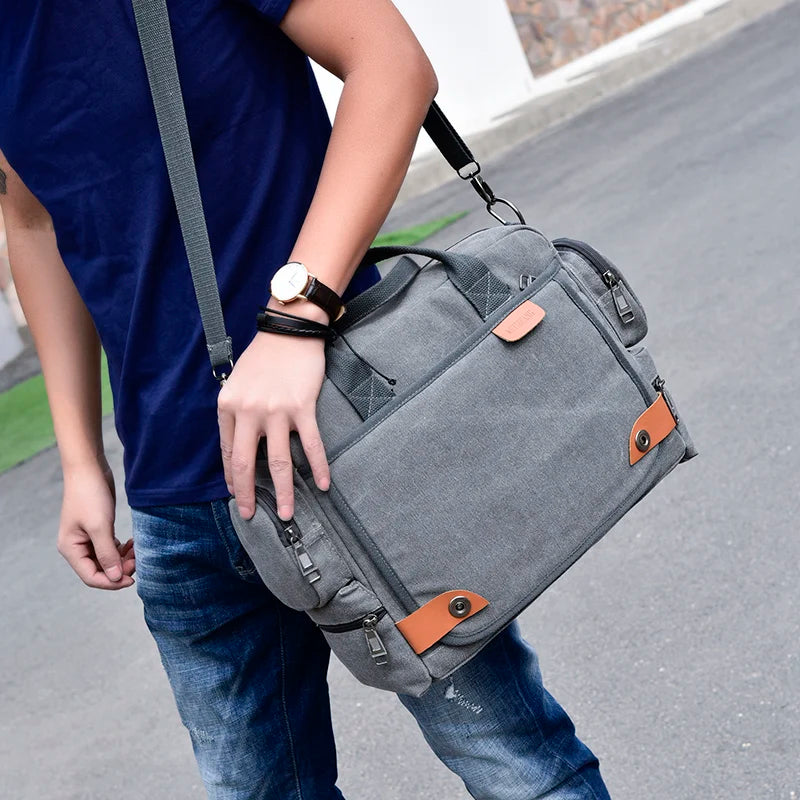 A man wearing a blue shirt and jeans is holding a gray canvas crossbody messenger bag with black straps.