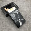 A black cigarette case with a silver sword design and a gold-colored cigarette inside, resting on a gray fabric surface.