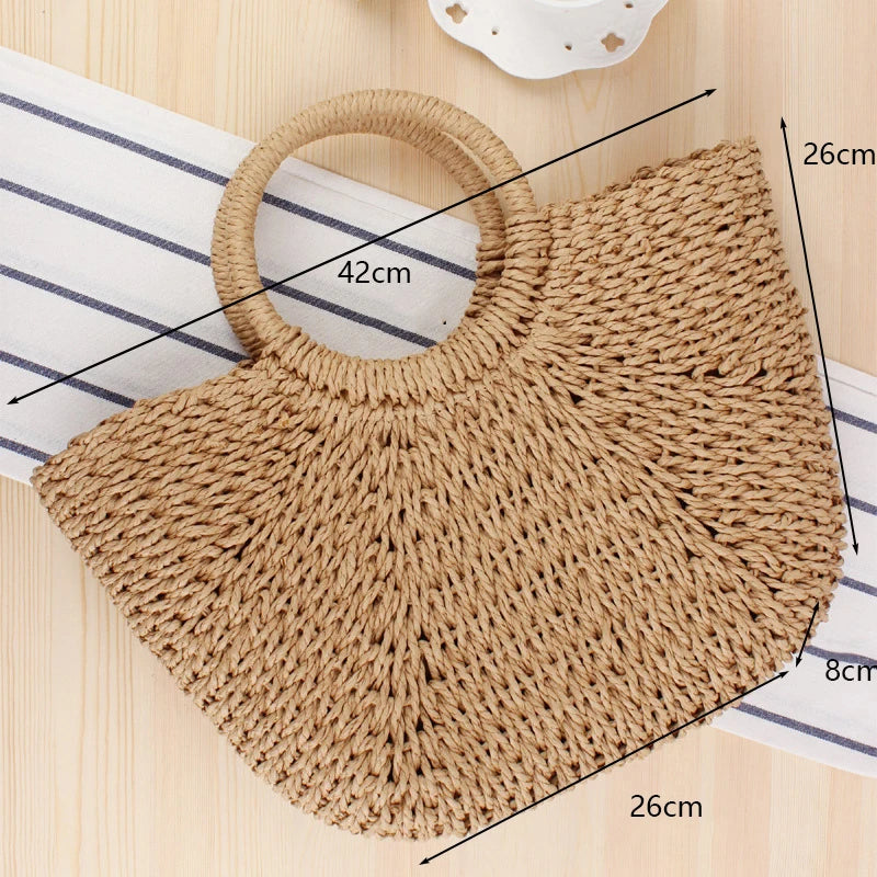 A handwoven straw moon bag with a round top handle shown on a wooden surface.