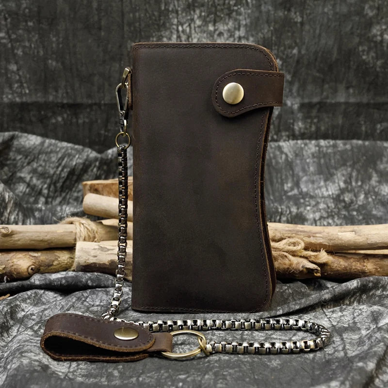 A dark brown genuine leather bifold wallet with a leather strap and metal chain resting on a dark fabric.