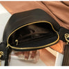 A black PU leather crossbody sling bag with gold hardware is shown against a tan fabric background.