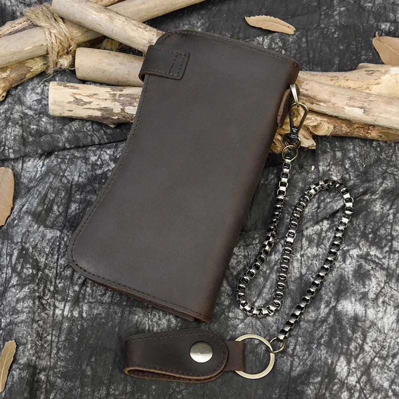 A dark brown genuine leather bifold wallet with a metal chain is displayed on a textured gray surface next to wooden sticks.